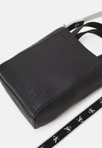 Borsa a tracolla nera in pelle compatta con logo CK in rilievo e tracolla regolabile con marchio su sfondo bianco.