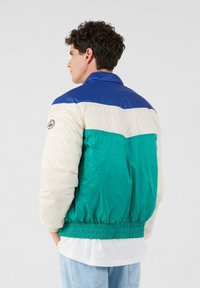 Uomo con capelli ricci che indossa un giubbotto colorato a blocchi blu, bianco e teal, in piedi di lato con le mani in tasca su uno sfondo semplice.