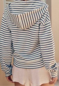 Pruhovaná mikina v krémové barvě s navy modrými pruhy, s kapucí na šňůrku a dlouhými rukávy. Tkanina vypadá m�ěkce a lehce.