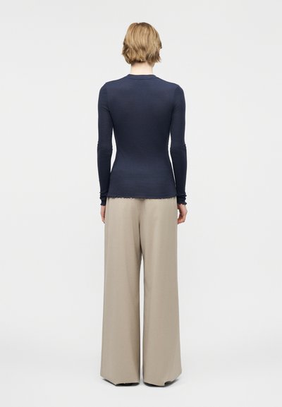 Άτομο με κοντά ξανθά μαλλιά, φοράει εφαρμοστή μπλε σκούρα μπλούζα με μακριά μανίκια και χαλαρό μπεζ παντελόνι με φαρδιά πόδια, στέκεται γυρισμένο πλάτη σε απλό φόντο.