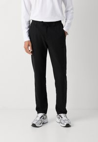 Zwarte lichtgewicht broek met een elastische tailleband en een verstelbaar trekkoord, gecombineerd met een witte longsleeve shirt en zilveren sportschoenen.