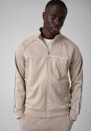 Mann trägt eine beige gemusterte Jacke mit Reißverschluss, weißen Streifen und einem Logo am Ärmel, steht vor einem schlichten grauen Hintergrund.
