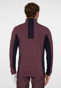 Lila långärmad tröja med svarta sidopaneler, hög krage och framträdande logotypdetalj på nackpartiet. Slät, stretchig tyg.