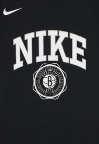 Juodas medvilninis marškinėliai su išsiskiriančiu baltu "NIKE" užrašu ir apvaliu Brooklyn Nets logotipu, kurį papildo krepšinio dizainas ir aplinkinis tekstas.