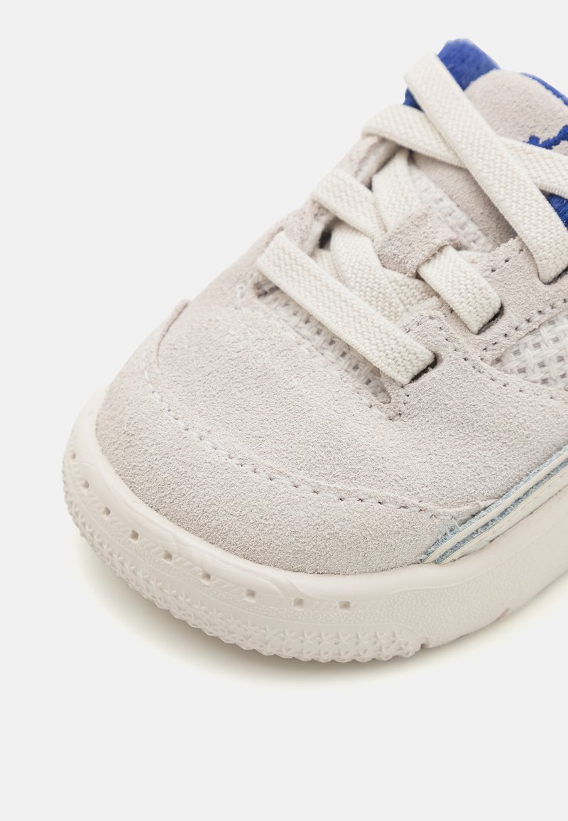 Zapatilla gris claro con parte superior de ante, suela de goma blanca, acentos azules y diseño de cordones. Superficie texturizada con detalle de malla en los laterales.