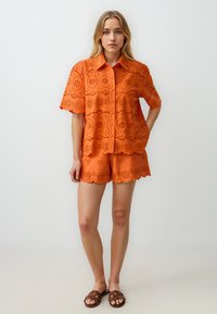 Oranje blouse met oogjes, korte mouwen en golvend zoom, gecombineerd met bijpassende shorts. Glad textuur, gedetailleerde bloemen uitsparingen en knopen aan de voorkant.