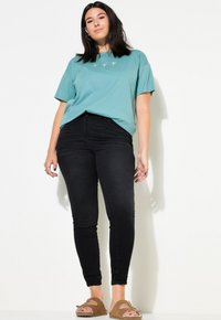 Camiseta de algodón en color azul verdoso con bordado sutil, combinada con jeans ajustados de cintura alta en color negro y zapatos de slip-on marrones. Texturas suaves en toda la prenda.