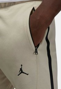 Beiga träningsbyxor med en svart Jordan-logotyp, sidofickor med dragkedja och svarta sidolinjer. Tillverkade av mjukt, elastiskt tyg.