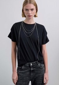 Camiseta negra de manga corta con textura suave, combinada con jeans gris oscuro. Tres cadenas plateadas en capas realzan el escote.