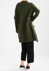 Pull en maille vert, long et surdimensionné, avec une finition texturée, associé à un pantalon noir court et des chaussures noires à enfiler.