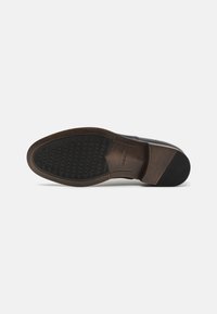 Zapato de cuero marrón con una parte superior lisa, una suela de goma oscura en contraste que presenta perforaciones circulares para la transpirabilidad y el agarre.