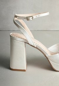 Biela sandála na vysokom hrubom podpätku s tenkými remienkami okolo členku a hladkým saténovým povrchom. Jemné prešitia a zaoblený tvar špičky.