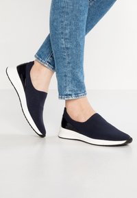 Marina slip-on sneakers med en texturerad tygövre, vit gummisula och blanka svarta detaljer på hälen för kontrast.