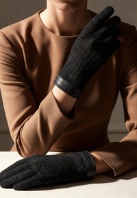 Gants en laine noirs avec une finition texturée et des poignets en cuir noir, portés avec un vêtement à manches longues marron, reposant sur une surface claire.