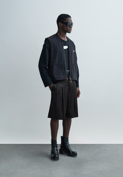 Μαύρο τζιν γιλέκο πάνω από μαύρο πουκάμισο με μακριά μανίκια, συνδυασμένο με σκούρα σορτς και γυαλιστερά μαύρα μποτάκια μέχρι τον αστράγαλο. Περιλαμβάνει ετικέτα στο πουκάμισο.