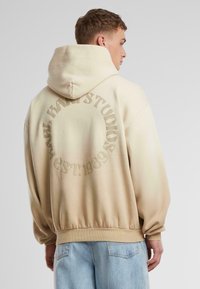 Beige oversized hoodie med dragsko på huvan, med en präglad cirkulär logotyp på baksidan; mjuk textur och ribbade ärmslut.