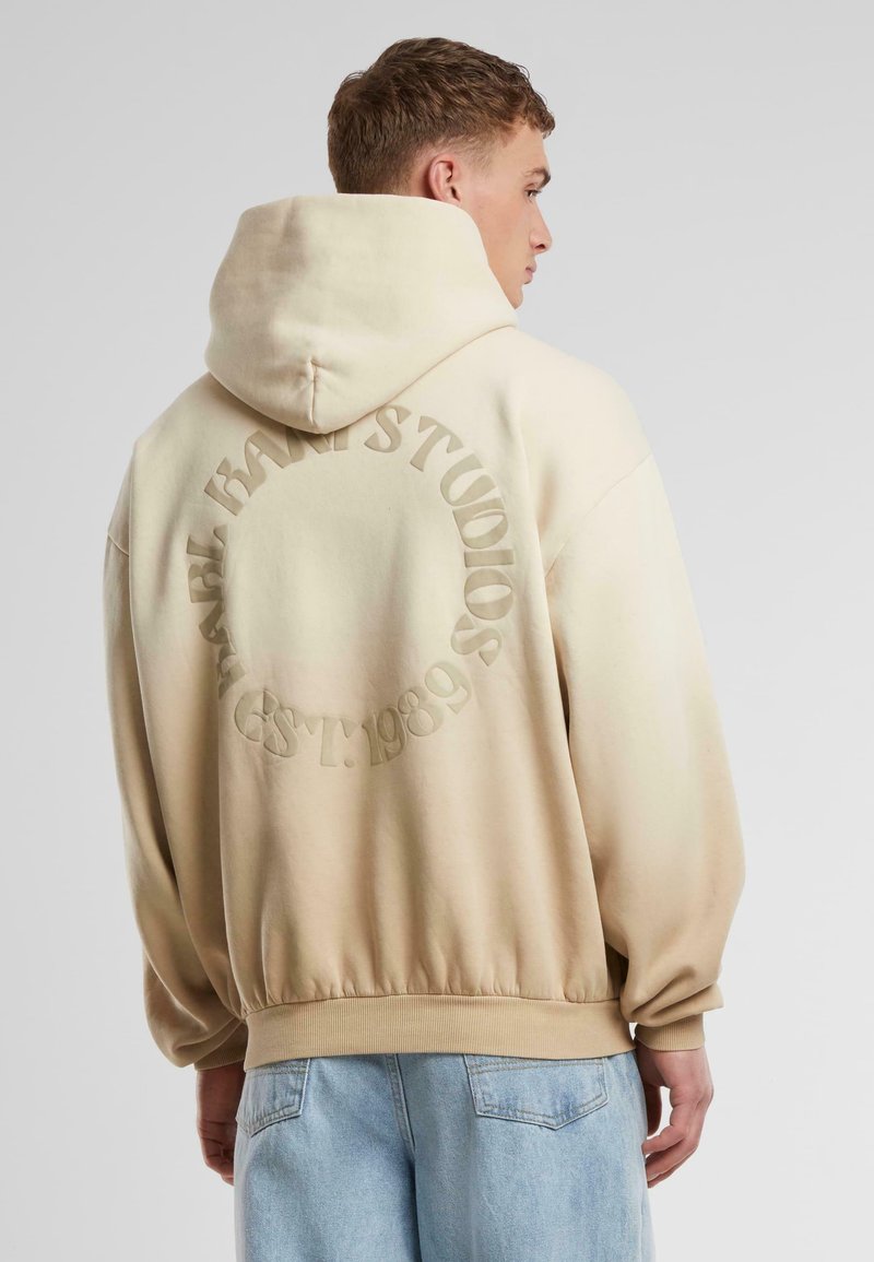 Beige oversized hoodie med dragsko på huvan, med en präglad cirkulär logotyp på baksidan; mjuk textur och ribbade ärmslut.