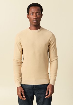 Man in een beige gebreide trui met lange mouwen en donkere spijkerbroek, staand tegen een lichtbeige achtergrond.