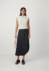 White cable-knit sleeveless top paired with a black, flowy midi skirt. Model carries a small beige handbag and wears black slide sandals.