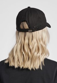 Zwarte pet gemaakt van stof, met een verstelbare band aan de achterkant en een gebogen rand. Toont golvend haar op de achtergrond.