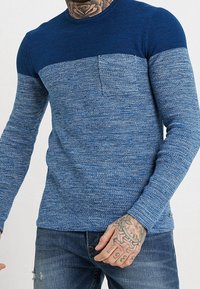 Blauer Strickpullover mit einem strukturierten Design, das einen einfarbigen dunkelblauen oberen Abschnitt und einen helleren blauen unteren Abschnitt aufweist. Enthält eine Brusttasche.