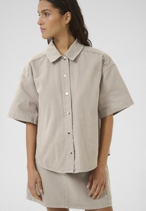 Camisa de mezclilla beige con mangas cortas, botones a presión en el frente y tachuelas decorativas en los hombros. Textura suave, corte relajado.