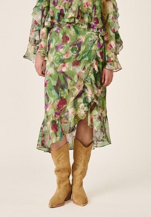 Falda midi verde con volantes y estampado floral combinada con botas beige de ante estilo vaquero hasta la rodilla sobre un fondo claro.