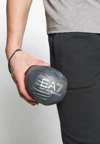 Šedá péřová bunda složená do kruhového tvaru, s bílým logem EA7 Emporio Armani. Hladký nylonový materiál, snadno přenosná.