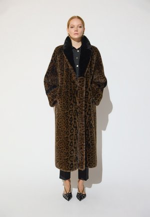 Mujer con un abrigo largo de piel marrón con estampado de leopardo con cuello negro, pantalones negros y tacones puntiagudos negros con tirantes de cadenas doradas.