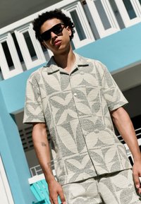 Koszula na krótki rękaw zapinana na guziki, wykonana z teksturowanej, jasnobeżowej tkaniny z geometrycznymi wzorami liści, w komplecie z dopasowanymi szortami.