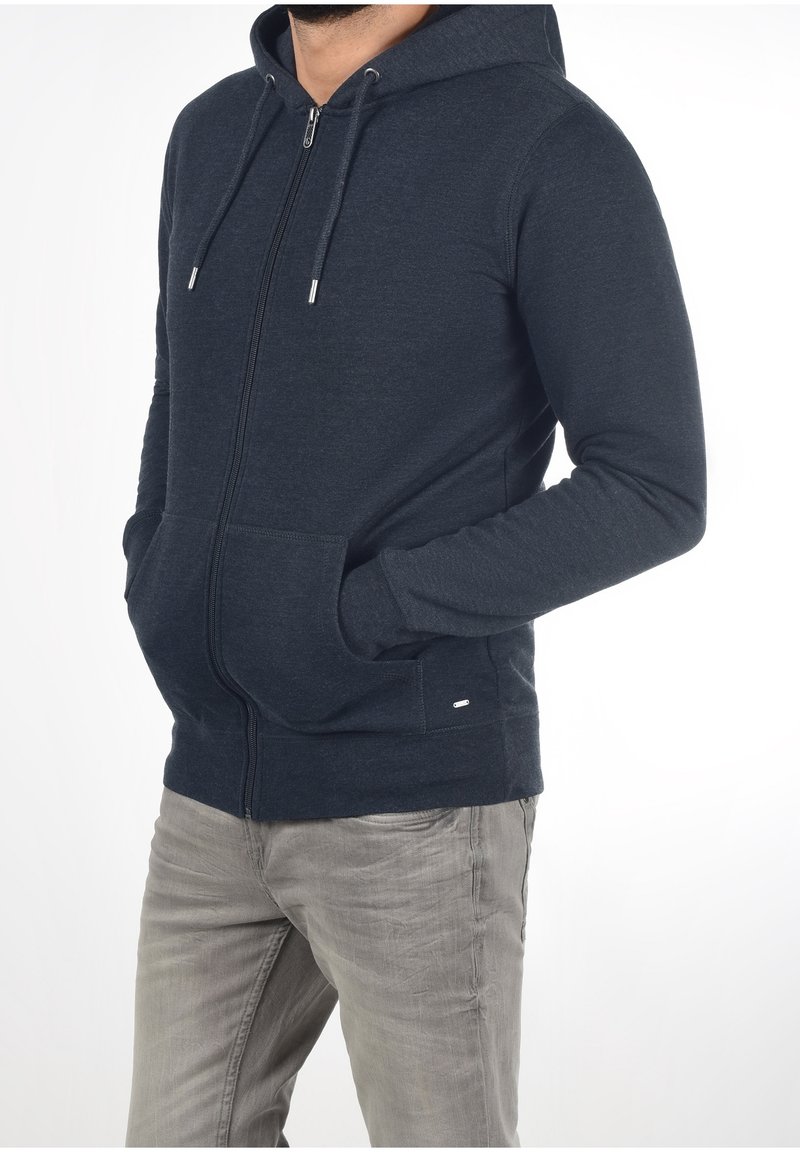 Homme portant un sweat à capuche zippé bleu marine foncé, les mains dans les poches avant, et un pantalon gris clair, debout devant un fond blanc.