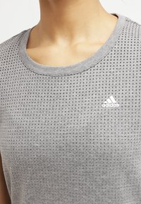 Zblízka osoba v šedém tričku Adidas s malými černými tečkami a bílým logem Adidas na prsou.