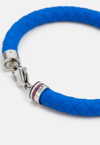 Pulsera de goma azul con un diseño trenzado, cierres de acero con una franja de acento roja y blanca, y un cierre tipo langosta.