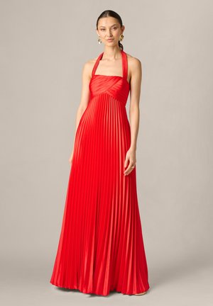 Femme portant une robe rouge plissée longue au sol avec des boucles d'oreilles dorées, debout devant un fond neutre uni.