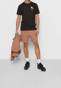 Svart bomullströja med en grön växtgrafik och orange etikett, i kombination med bruna shorts och svarta sneakers, håller en brun jacka.