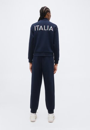 Osoba z warkoczami ubrana w granatowy dres z napisem "ITALIA" na plecach i białe trampki, stojąca na jednolitym tle.