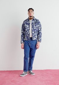 Chaqueta navy con estampado floral, camisa blanca, jeans azules y zapatillas gris. Tejido suave con un corte cuadrado y detalles de costura en contraste. Fondo rosa.