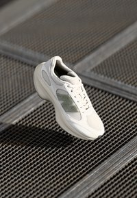 Baskets athlétiques blanches et grises suspendues au-dessus d'une surface métallique grillagée avec un motif géométrique en grille.