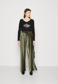 Top nero a maniche lunghe con logo decorato, abbinato a pantaloni a vita alta, verde oliva, plissettati e scarpe nere a punta.