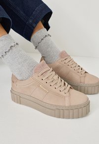 Beige sneakers with a textured upper and platform sole. Features flat laces and a subtle logo detail. Paired with gray frilled socks.