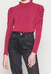 Red long-sleeve top with a ruffled high neckline and gathered detailing on shoulders, paired with black high-waisted jeans.