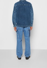 Giacca di jeans con lavaggio blu e maniche lunghe, abbinata a pantaloni cargo azzurri chiari con molte tasche e una vestibilità rilassata.