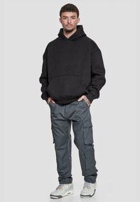 Svart oversized hettegenser med frontlomme, kombinert med grå cargo-bukser med flere lommer. Grå og hvite joggesko fullfører antrekket.
