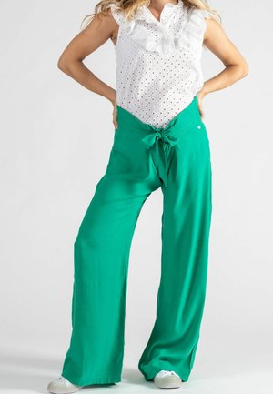 Pantalon large vert avec une ceinture à nouer à la taille, fabriqué en tissu léger. Assorti à un blouse blanche à volants et en dentelle.