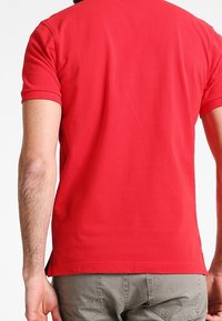 Homme portant un polo rouge uni à manches courtes et un pantalon gris, vu de dos sur fond blanc.