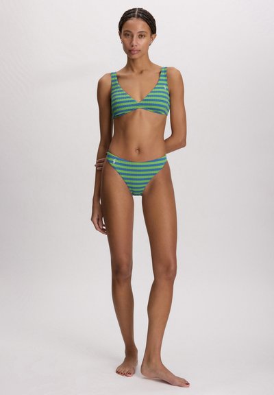 Γυναίκα που στέκεται ξυπόλητη φορώντας μαγιό με πράσινες και μπλε ρίγες, τοπ με V-λαιμόκοψη και σλιπ με ψηλή κοπή, μπροστά σε απλό φωτεινό φόντο.
