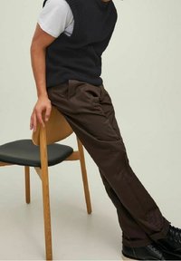Dunkelblauer Strick-Ärmelloser Pullover über einem weißen Hemd, braune Hosen mit Falten und schwarze Stiefel, sitzend auf einem Holzstuhl mit schwarzem Sitz.