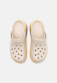Beige tresko laget av fleksibelt materiale med ventilasjonshull, forhøyet hæl, gul såleaccent og justerbar stropp bak.