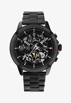 Montre chronographe noire avec un bracelet en acier inoxydable. Présente un cadran squelettique, des index argentés et une touche de rouge-blanc-bleu.