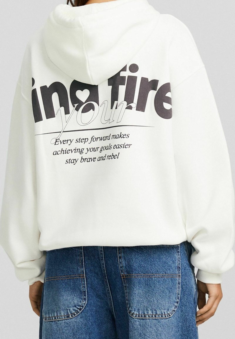 Personne portant un sweat à capuche blanc avec l'inscription "in fire your" et un texte motivant imprimé au dos, associée à un jean bleu en denim.